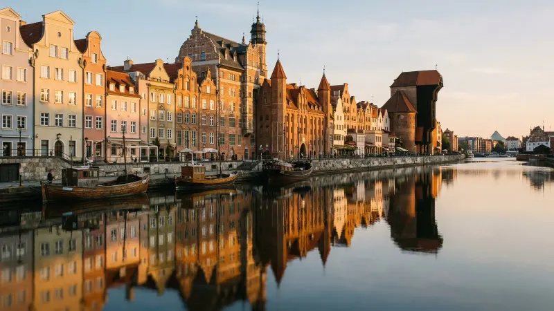 Golden-hour view of Gdańsk's brick and pastel riverfront houses reflected in the calm Motława river, with the old wooden port crane on the right.