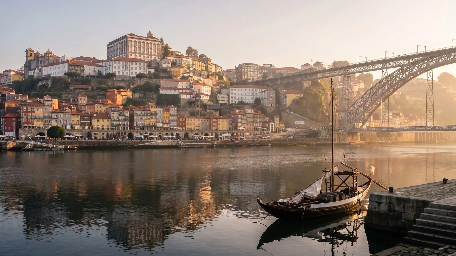 Porto's tiered riverfront houses glow in golden evening light above the Douro, with a double-deck iron bridge arching across the calm water.
