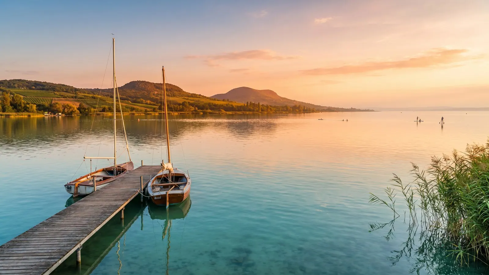 Golden-hour view across the calm, shallow waters of Lake Balaton toward the low volcanic hills and vineyards of the Hungarian shore, with a wooden jetty and distant sailboats.