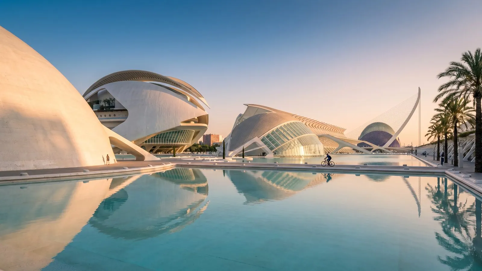The white, curved shells of Valencia's City of Arts and Sciences reflected in a calm turquoise pool under a warm golden-hour sky.