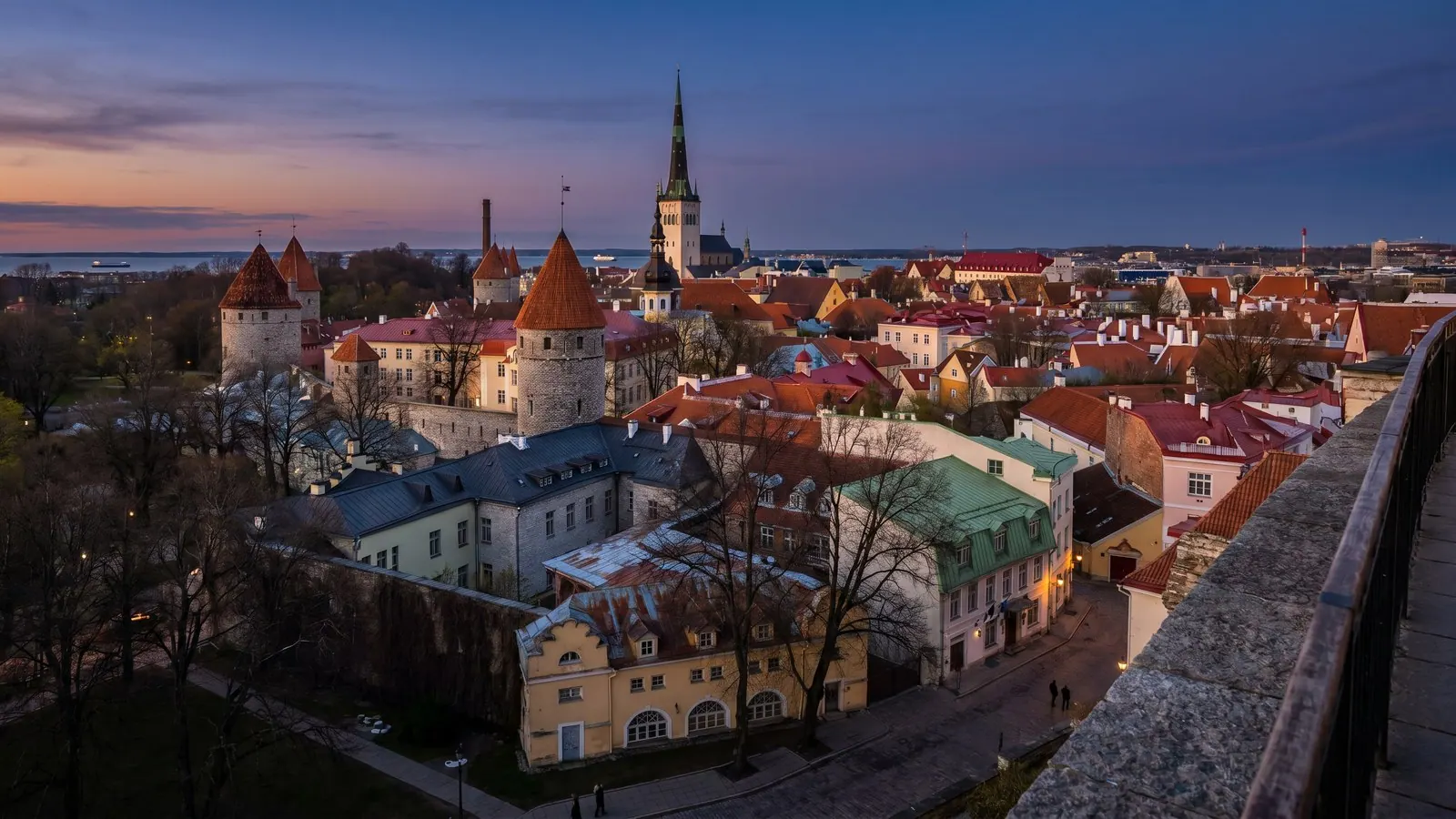 Red-roofed medieval towers and church spires of Tallinn's Old Town glowing under a soft golden-blue summer dusk sky, with the Baltic Sea beyond.