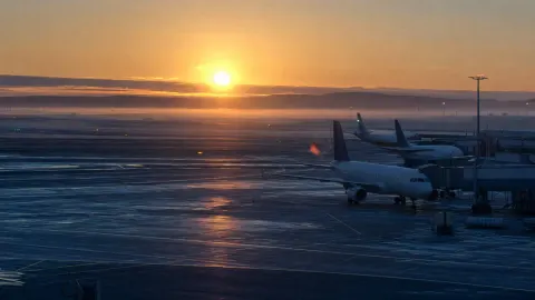 Parked passenger jets on a calm airport apron at dawn under a soft golden sky with light mist over the tarmac.