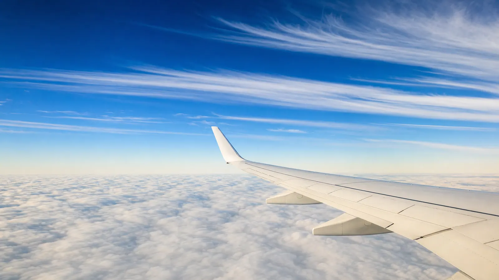 The plain white wing of an airliner at cruising altitude above a flat cloud deck, with long parallel bands of high cirrus streaked across a deep blue autumn sky.